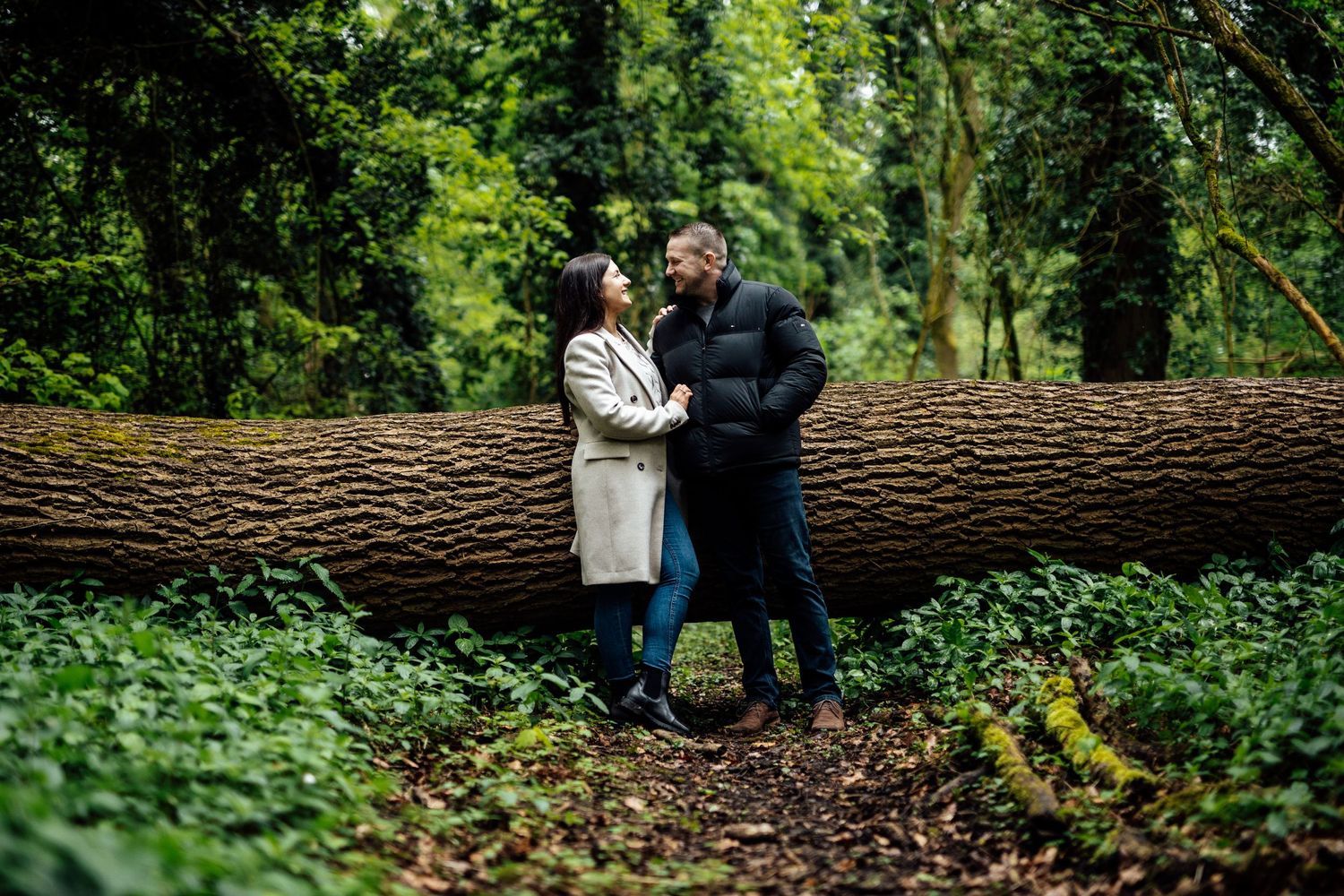 A couple shares a tender moment while standing on a fallen tree trunk in a lush forest.