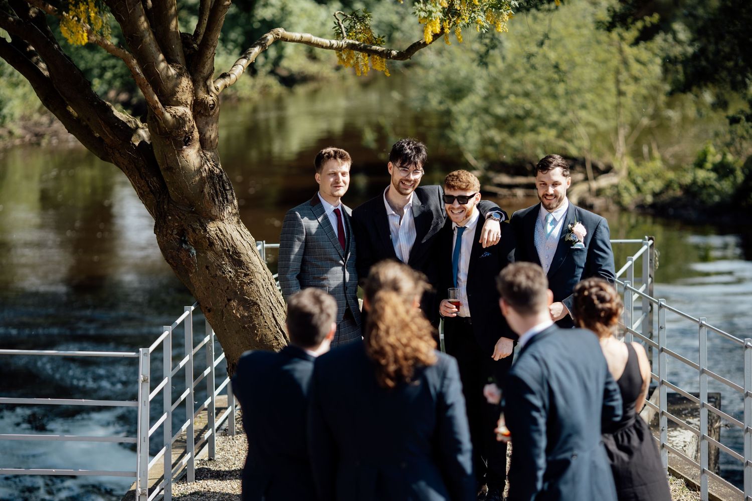 Wedding party gathers around tree by waterfront with metal railing.