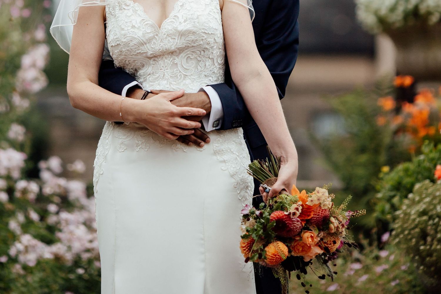 Close-up of couple's hands with wedding rings and orange floral bouquet against garden backdrop.