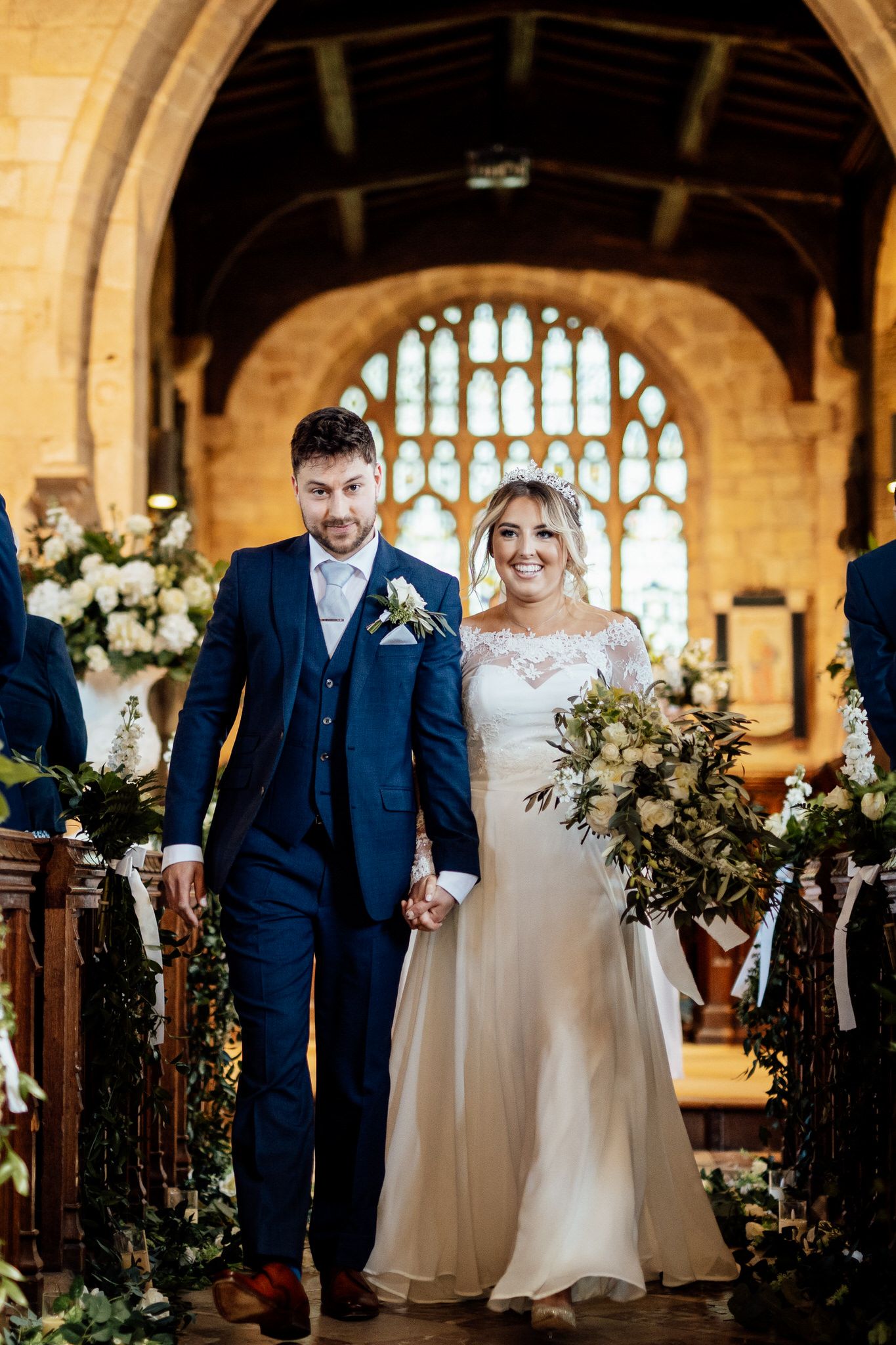 Couple walking down church aisle decorated with white flowers and greenery.