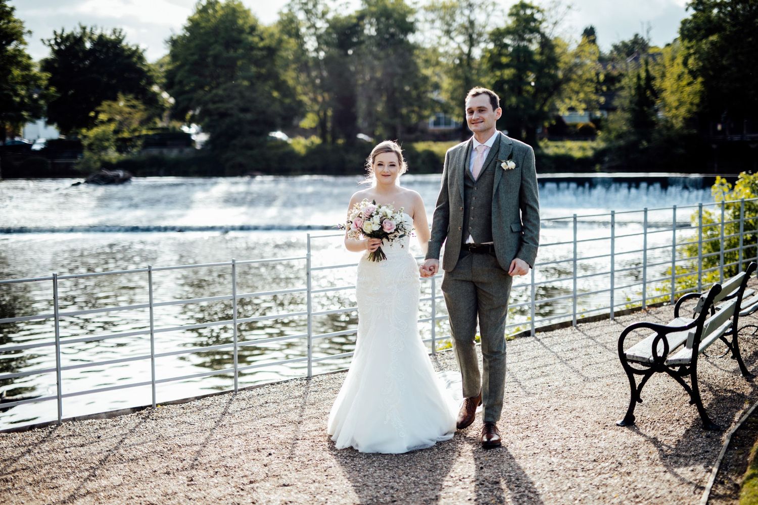 Couple poses for wedding portrait by waterfront with fountain and trees in background.