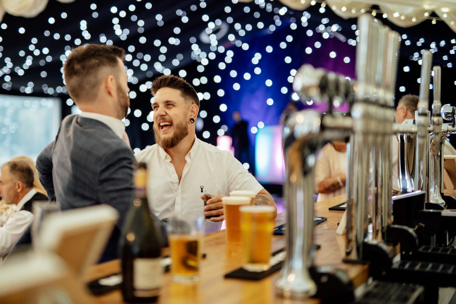 Wedding reception guests enjoying drinks and conversation at a festively decorated bar with bokeh lights.