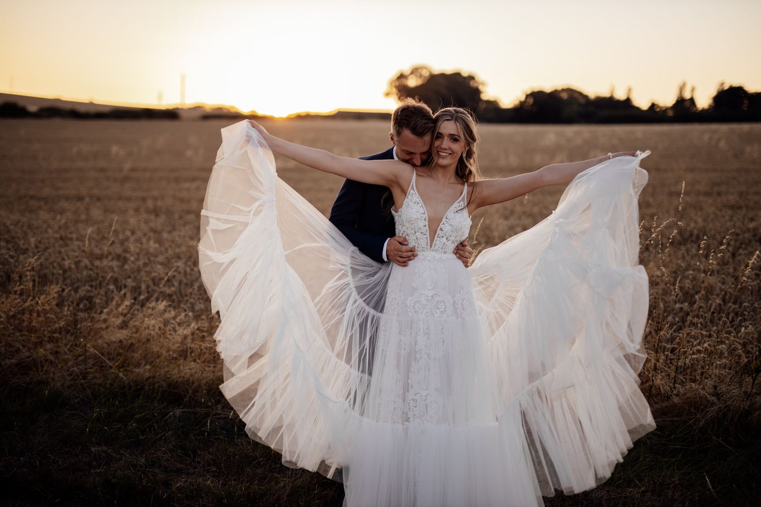 A bride twirls her flowing white dress in a golden field at sunset while being embraced.