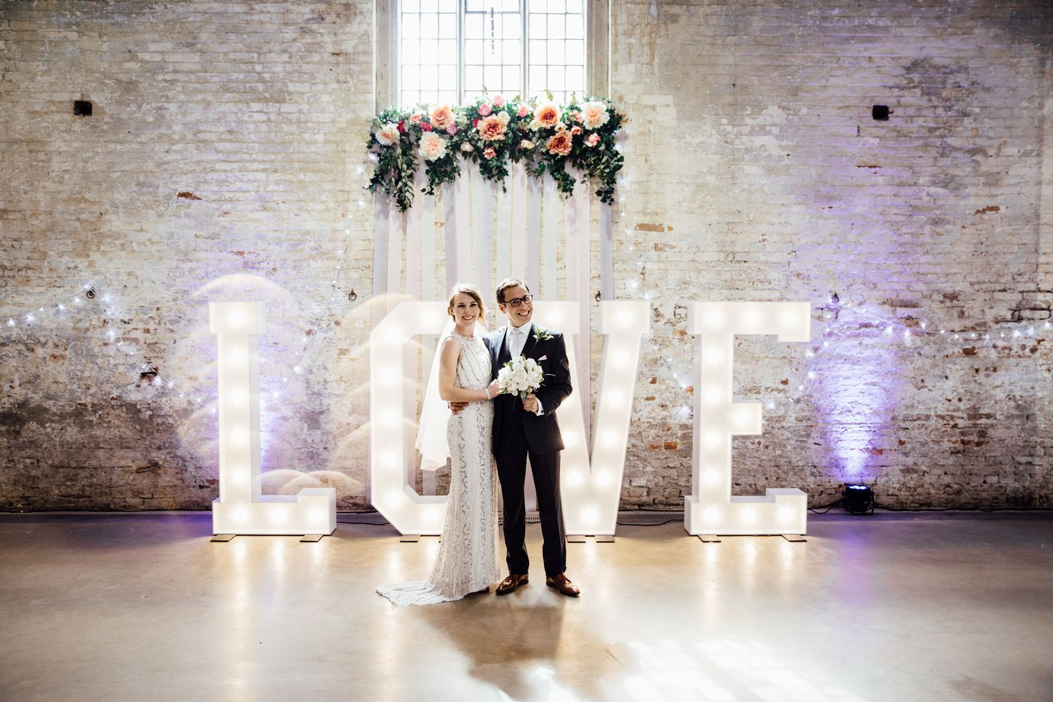 A couple stands in front of illuminated LOVE letters with floral decor in a rustic brick venue.