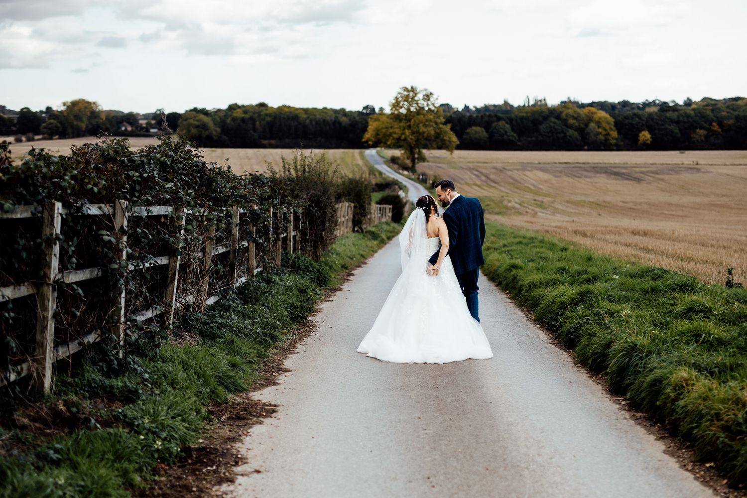 Couple walking through a scenic rural path lined with trees and fields on their wedding day.