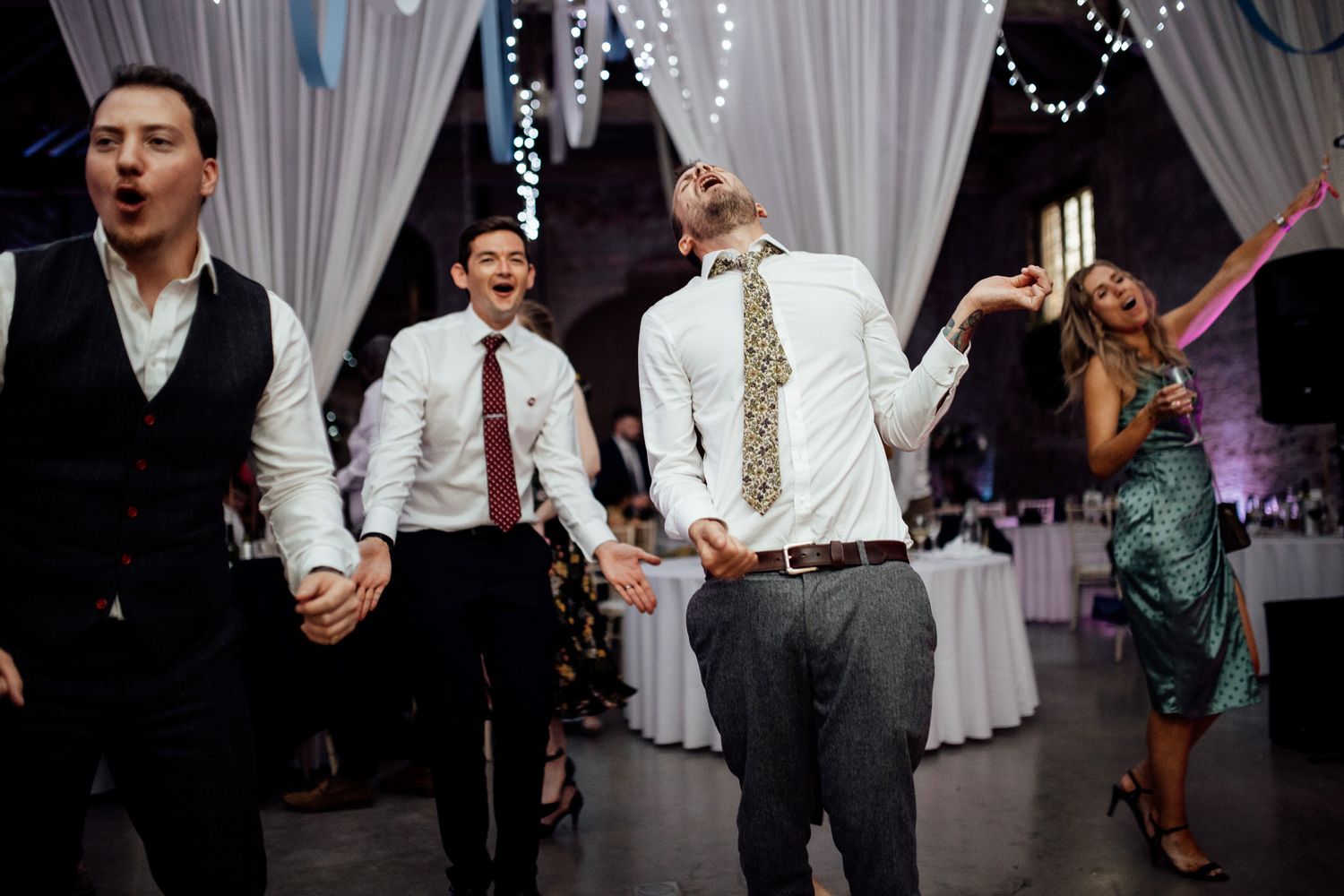Wedding guests dancing and celebrating on a dance floor decorated with string lights and white drapes.