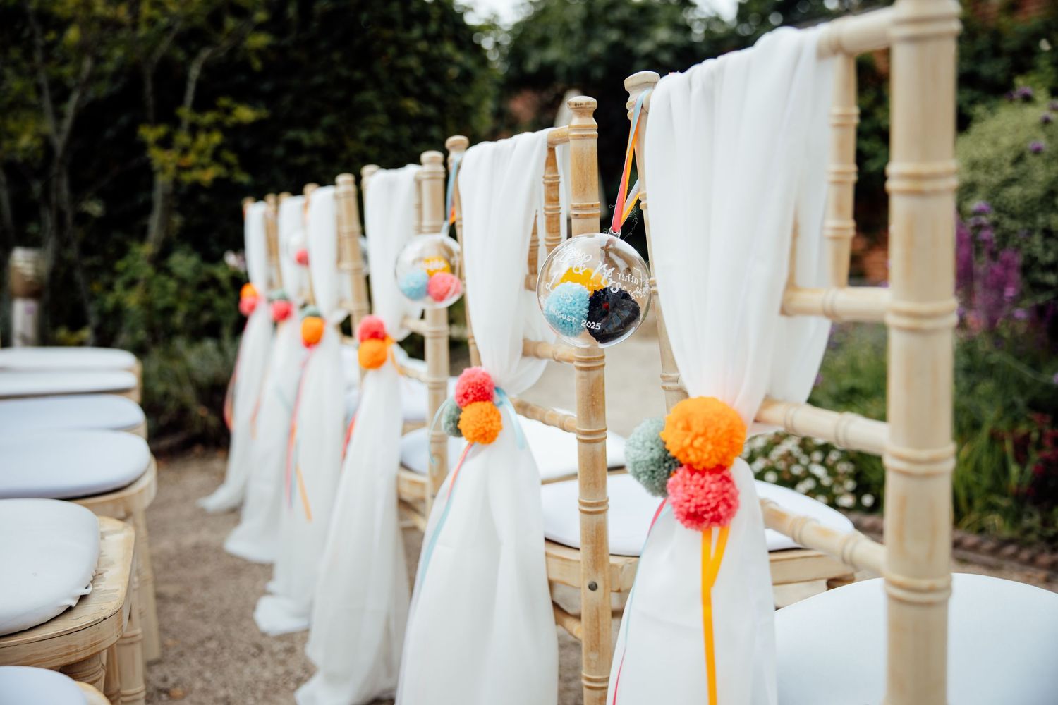 White fabric draped ceremony chairs decorated with orange pom poms along garden path.