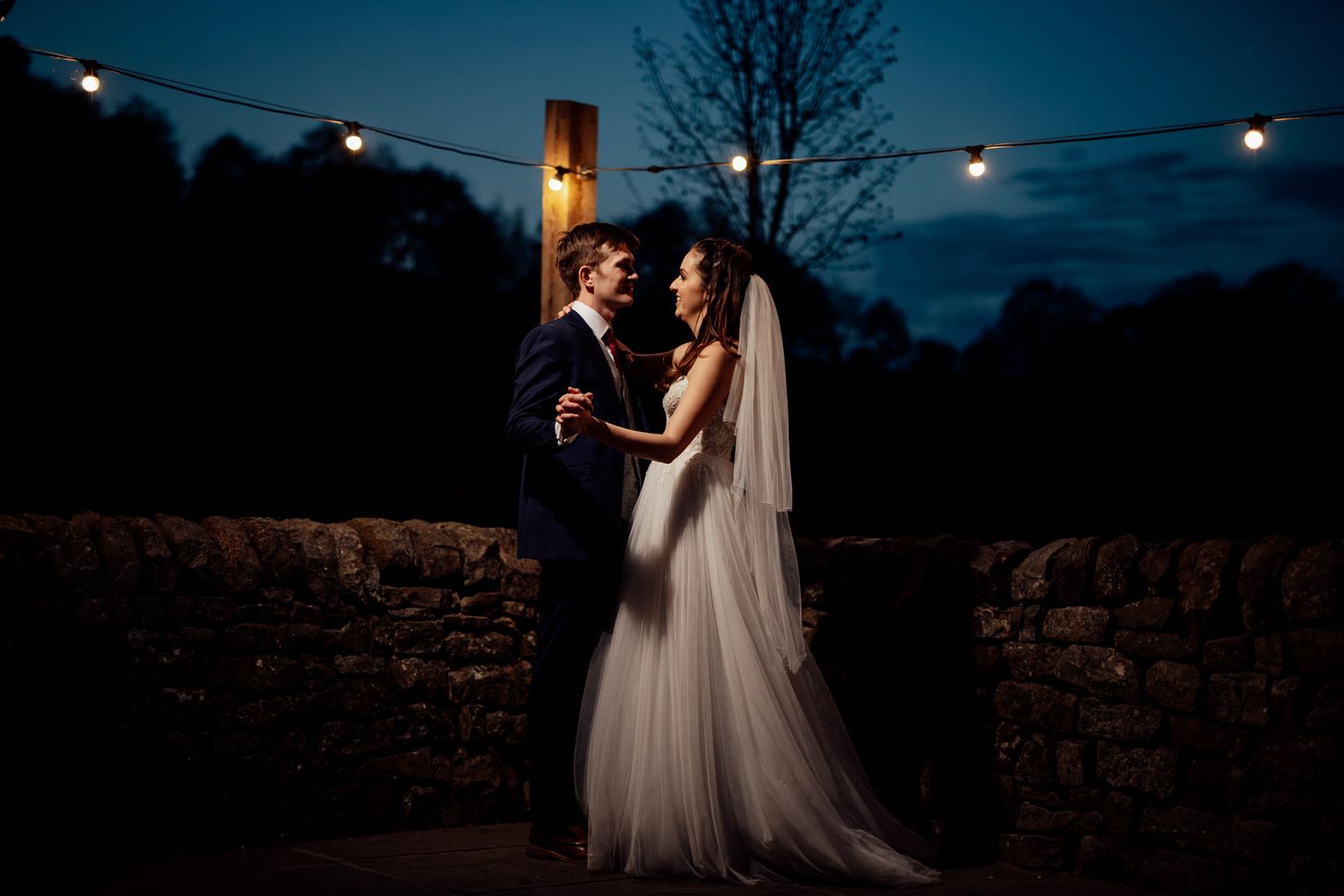 Evening wedding reception under string lights with stone wall backdrop.