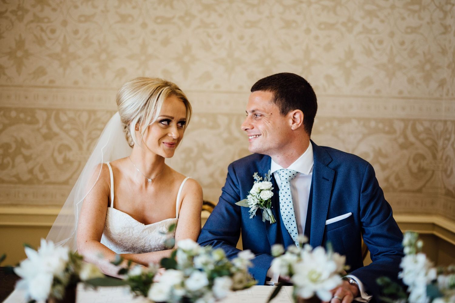 Wedding reception scene in an elegant room with ornate wallpaper and floral centerpieces.