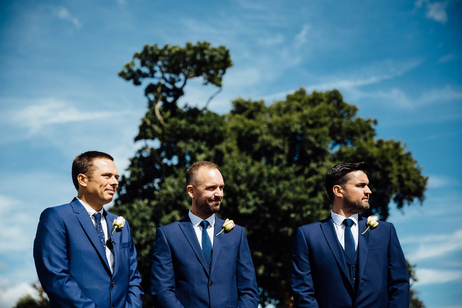 Three groomsmen in matching navy suits stand against a bright blue sky.
