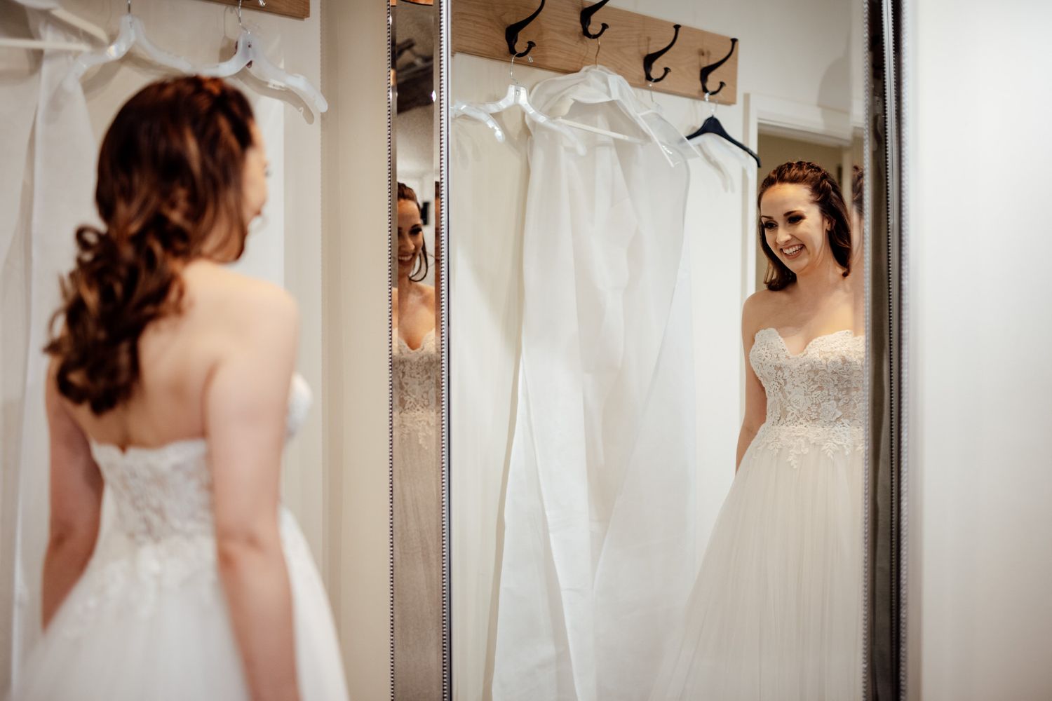 Bride looking at reflection in dressing room mirror with wedding gowns hanging.