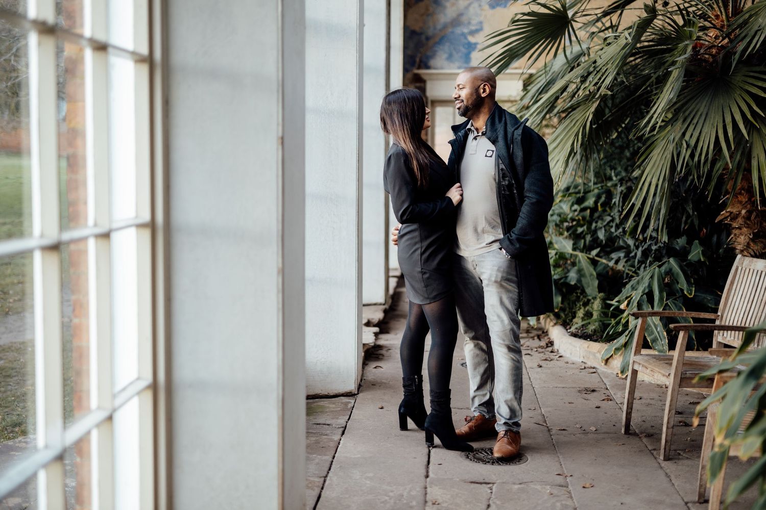 A couple in formal attire poses together on an outdoor patio with tropical plants and white columns.