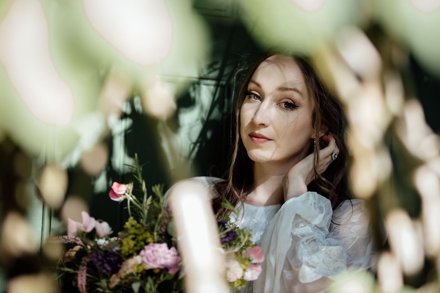 A person in a white lace dress poses outdoors with dreamy bokeh lighting and natural foliage in the background.