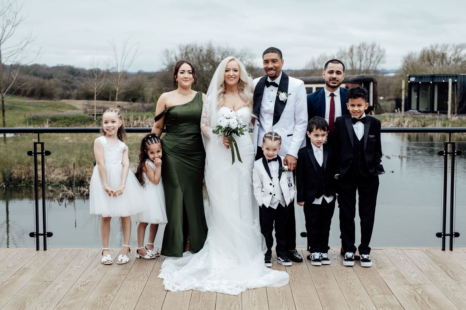Wedding party group photo with bride, groom, and children posing by a lakeside venue.