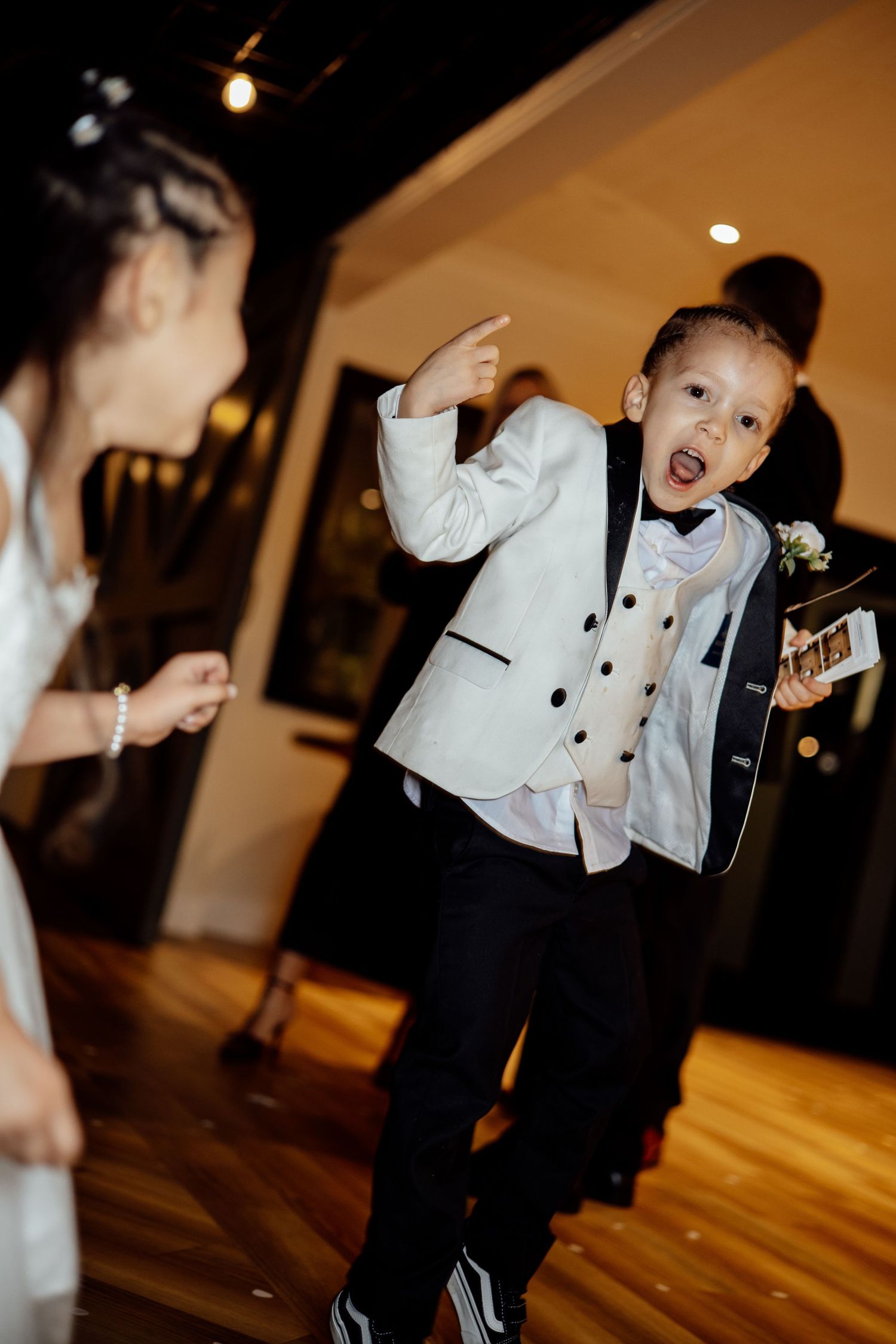 Excited young ring bearer in white tuxedo jacket pointing and dancing at wedding reception.