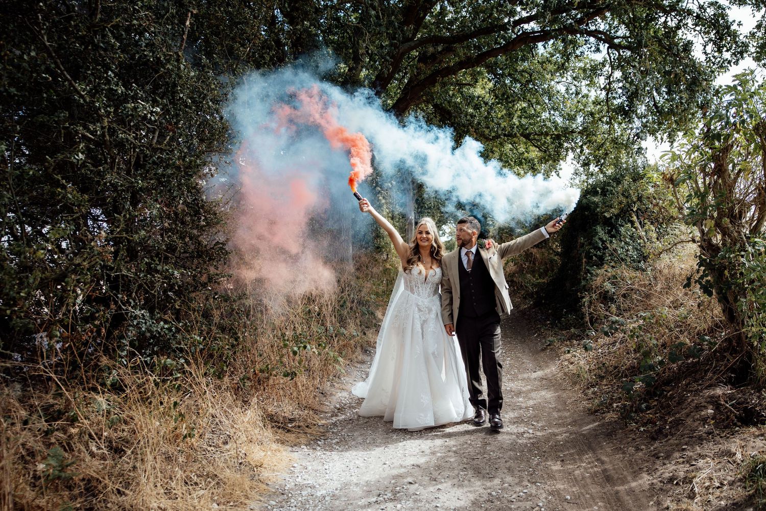 Bride and groom celebrate with colorful smoke bombs on a woodland path.