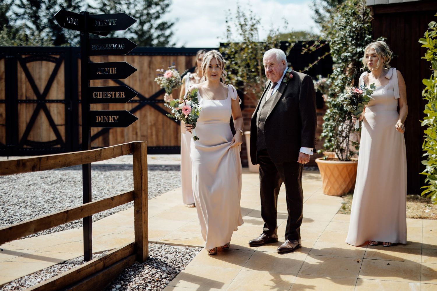 Bride in elegant gown walks with older gentleman and bridesmaid through rustic barn wedding venue gates.