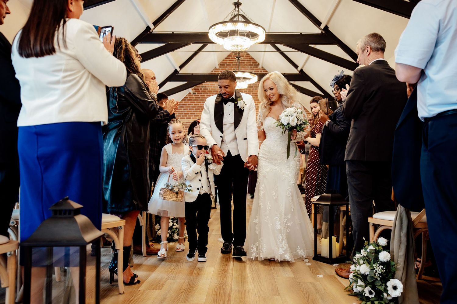 Bride and groom walking down the aisle with a flower girl as guests celebrate with confetti.