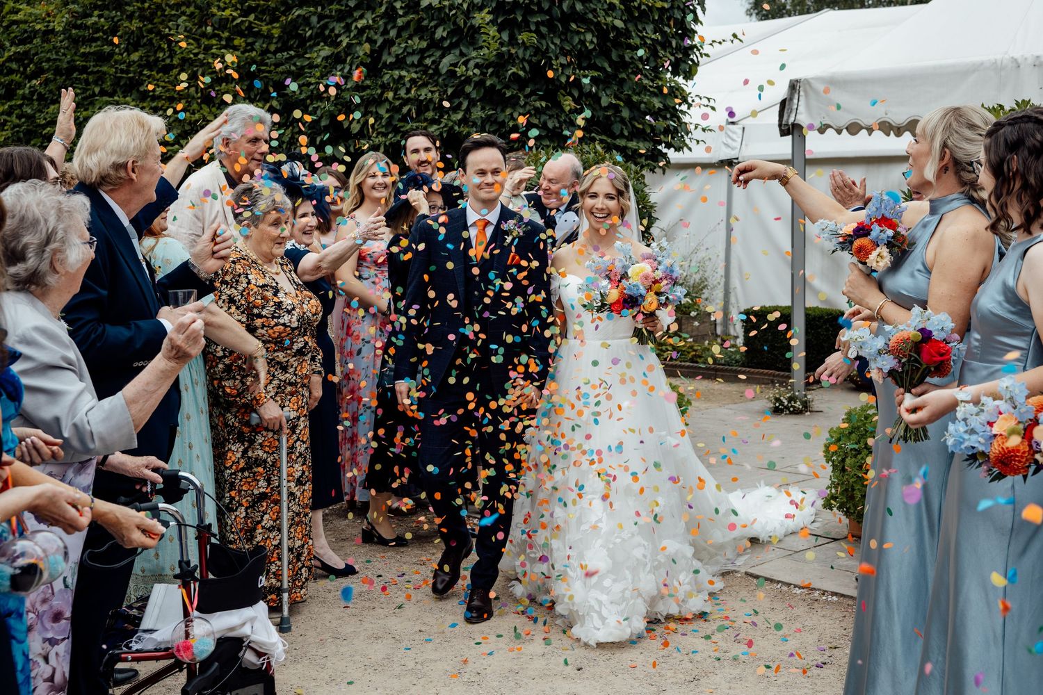 Wedding couple walks through shower of colorful confetti as guests celebrate along pathway at outdoor ceremony.