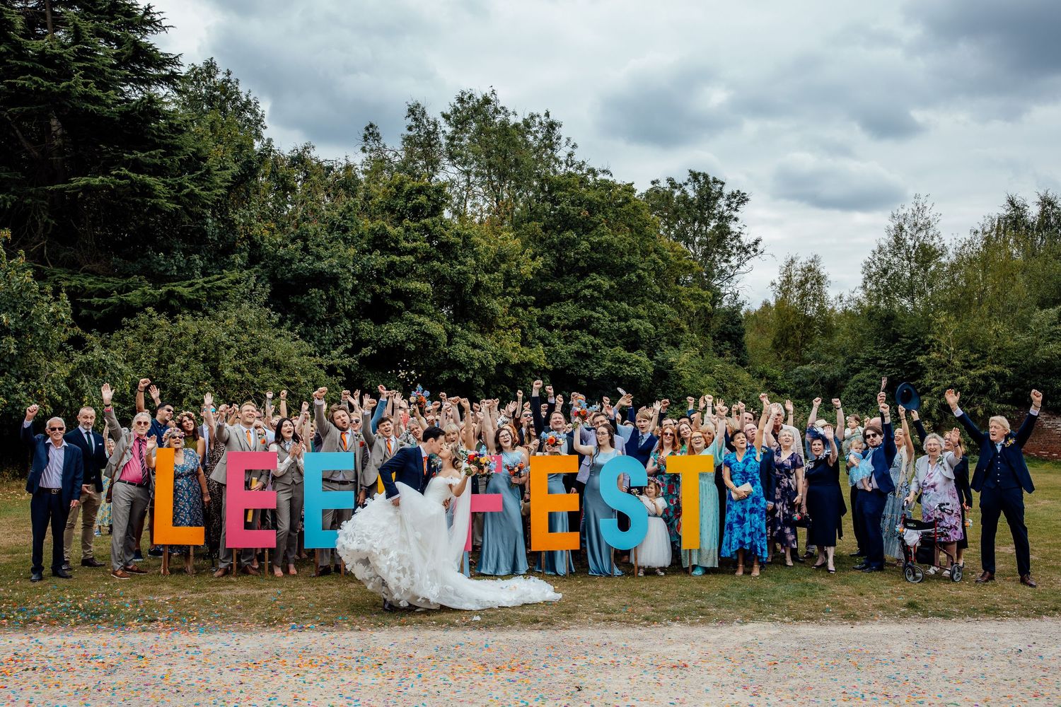 Large group wedding photo with guests arranged outdoors under cloudy skies.
