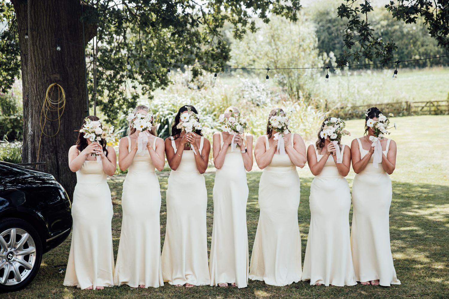 Bridesmaids in white dresses pose in line against natural outdoor backdrop.