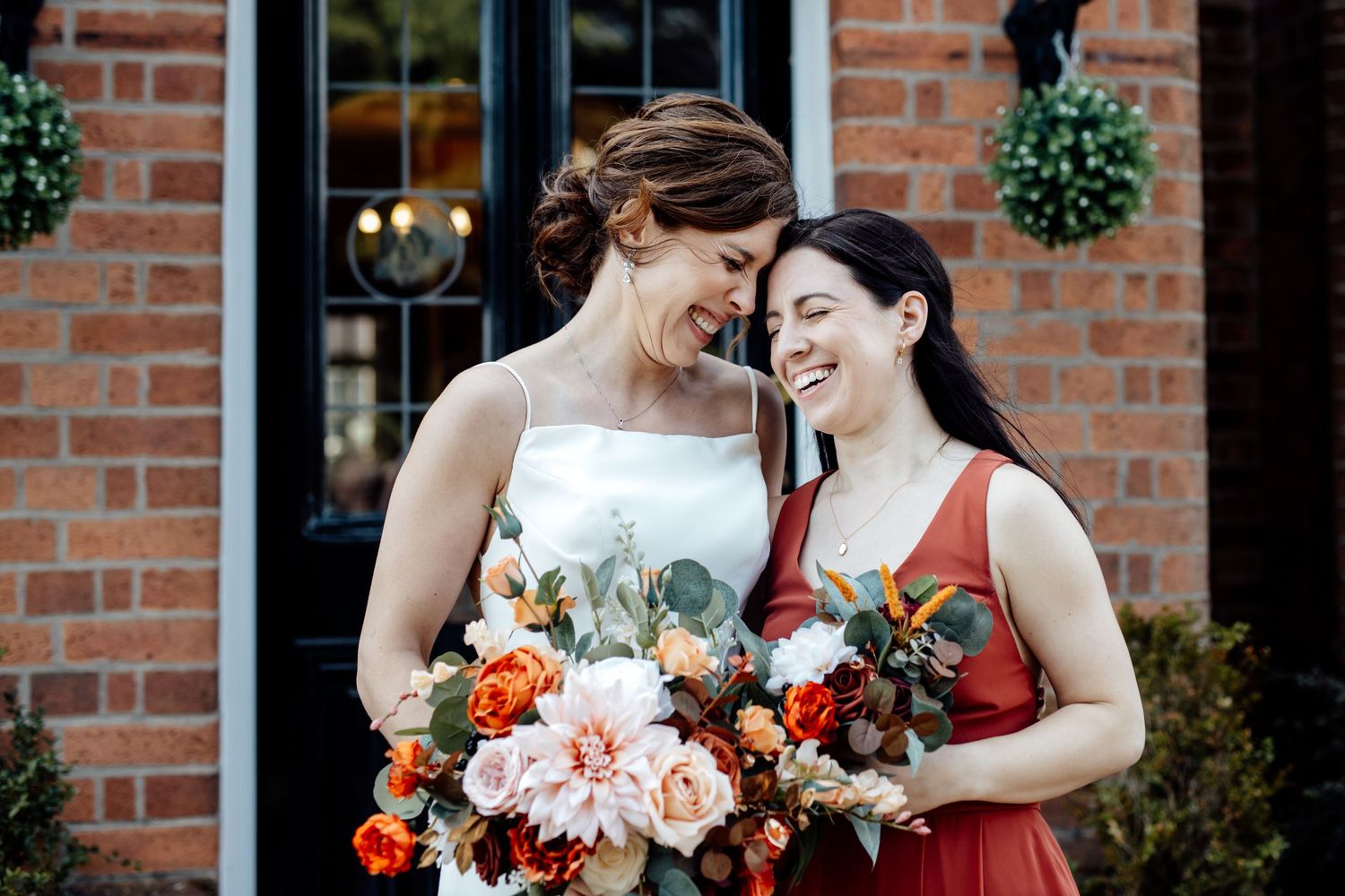 Bridesmaids in red dresses holding floral bouquets against brick wall.