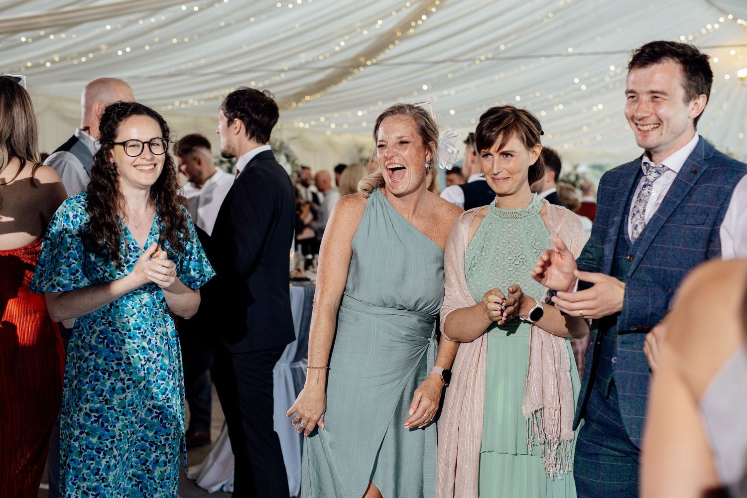 Wedding guests laugh and celebrate under twinkling lights at an indoor reception.