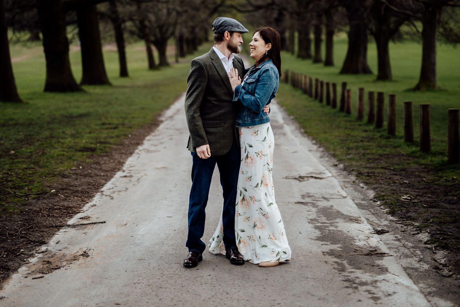 A romantic couple embraces on a tree-lined path wearing coordinated formal attire with casual outerwear.