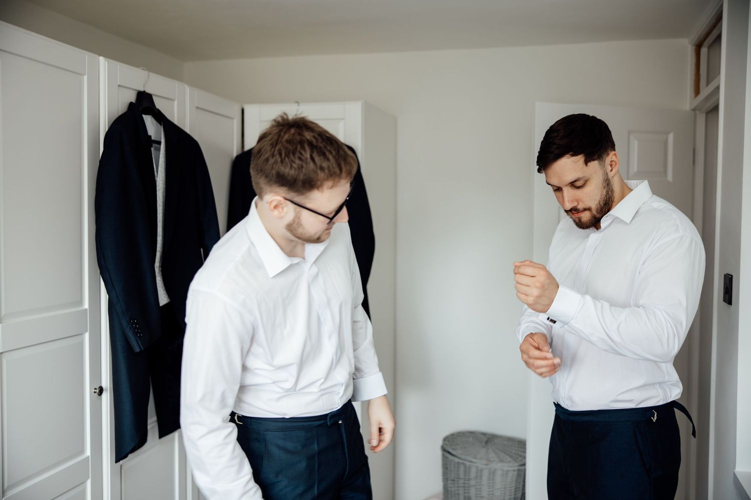 Two men in white shirts getting ready in a room with a jacket hanging on the wall.
