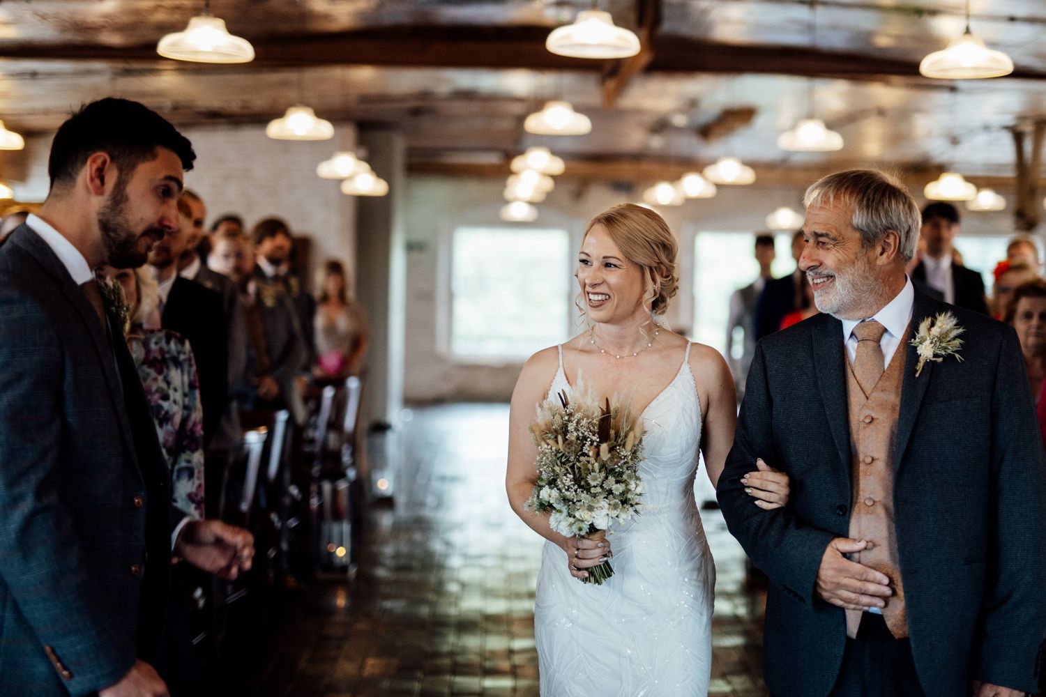 Wedding reception guests mingle in a rustic indoor venue with pendant lighting and wooden beams.