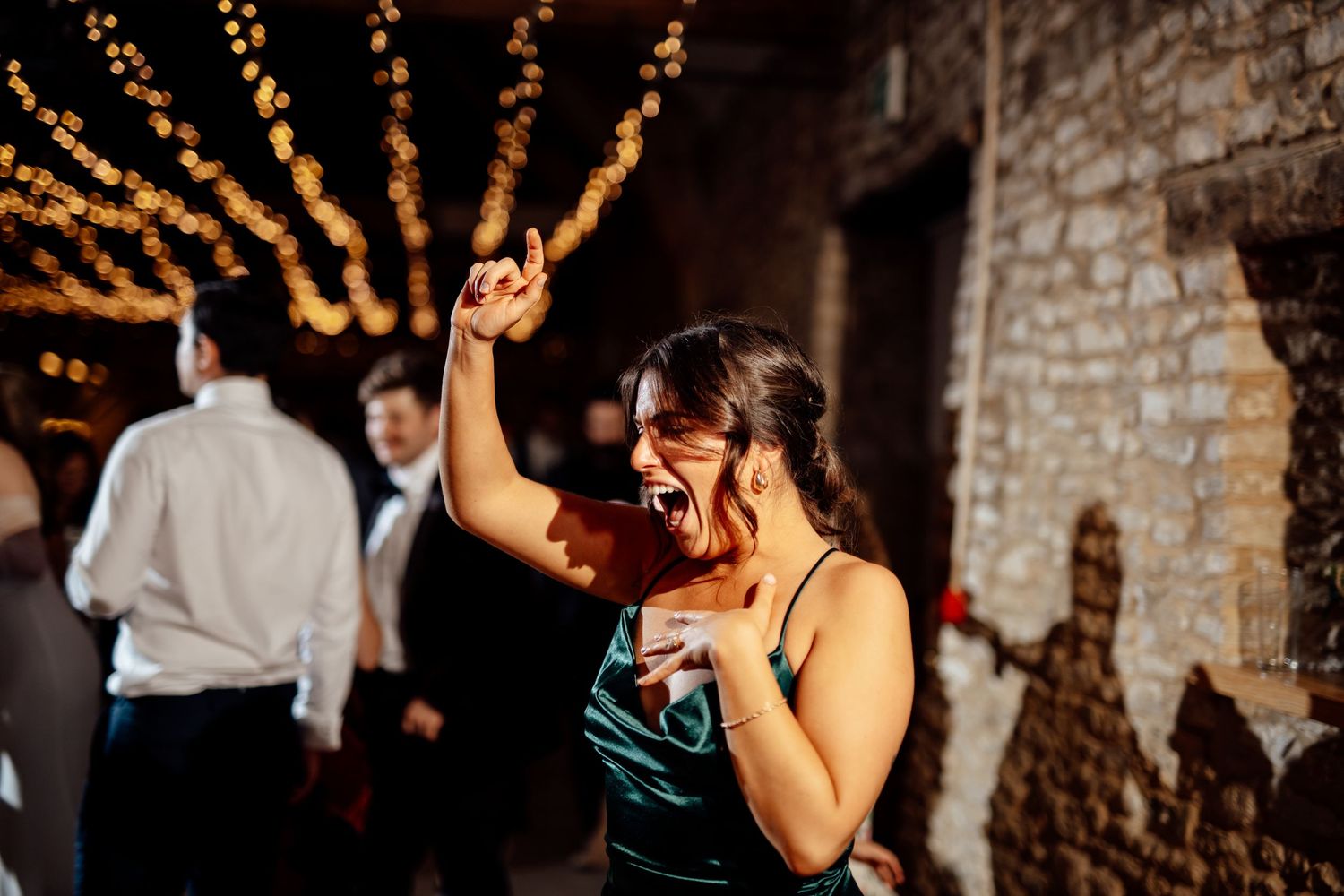 Warm glowing string lights illuminate stone walls at evening wedding reception.