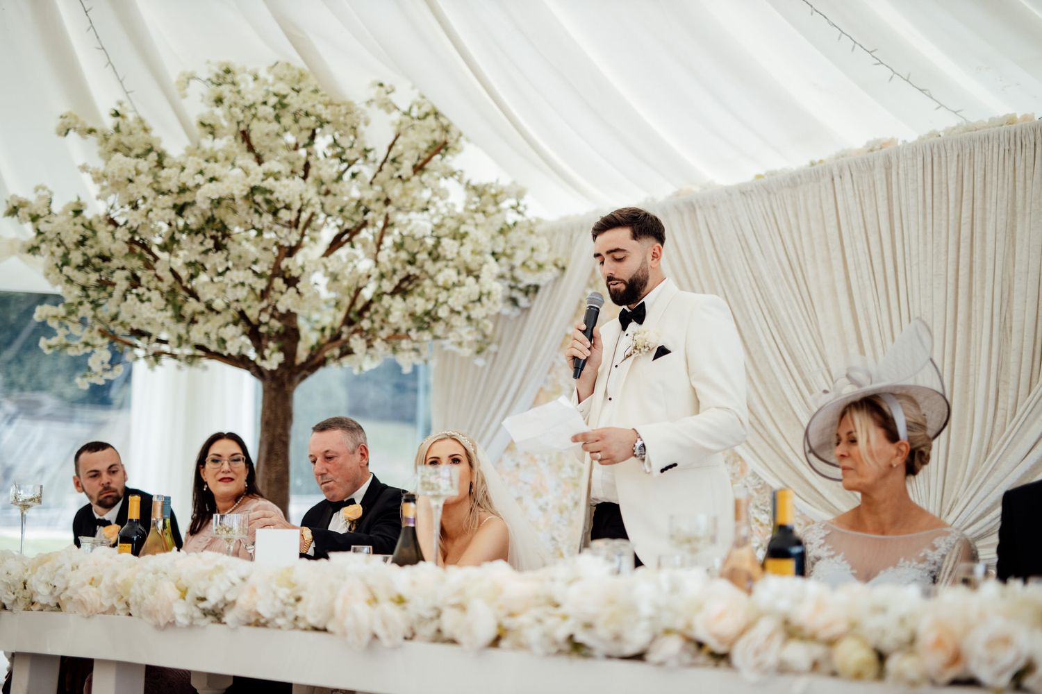 White draped reception tent decorated with elegant floral arrangements and crystal chandeliers.