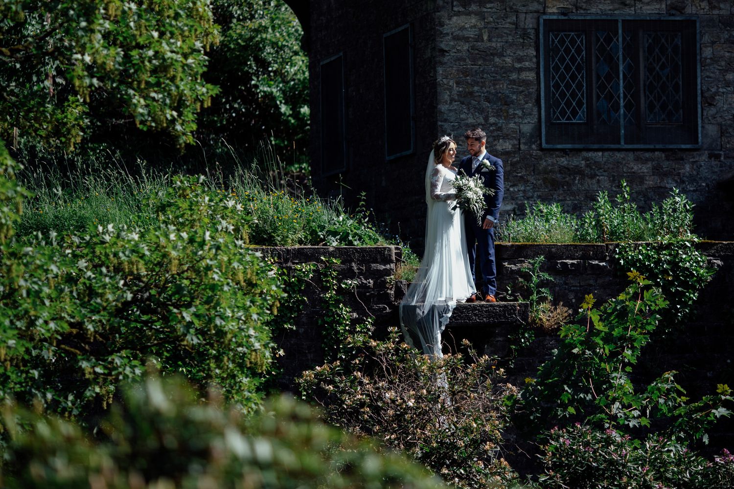 Romantic garden wedding portrait with stone wall and lush greenery.