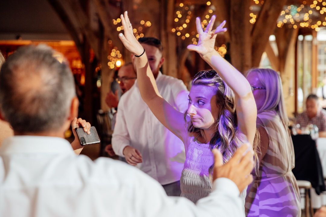 Wedding reception dance floor filled with joyful guests under string lights and purple uplighting.
