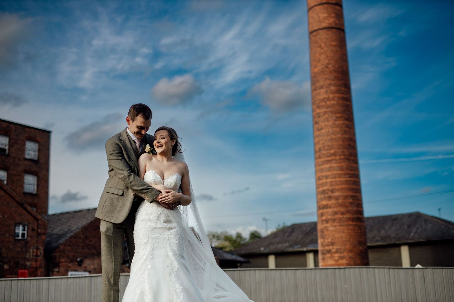Newlyweds pose together on rooftop with industrial brick chimney and blue sky backdrop.
