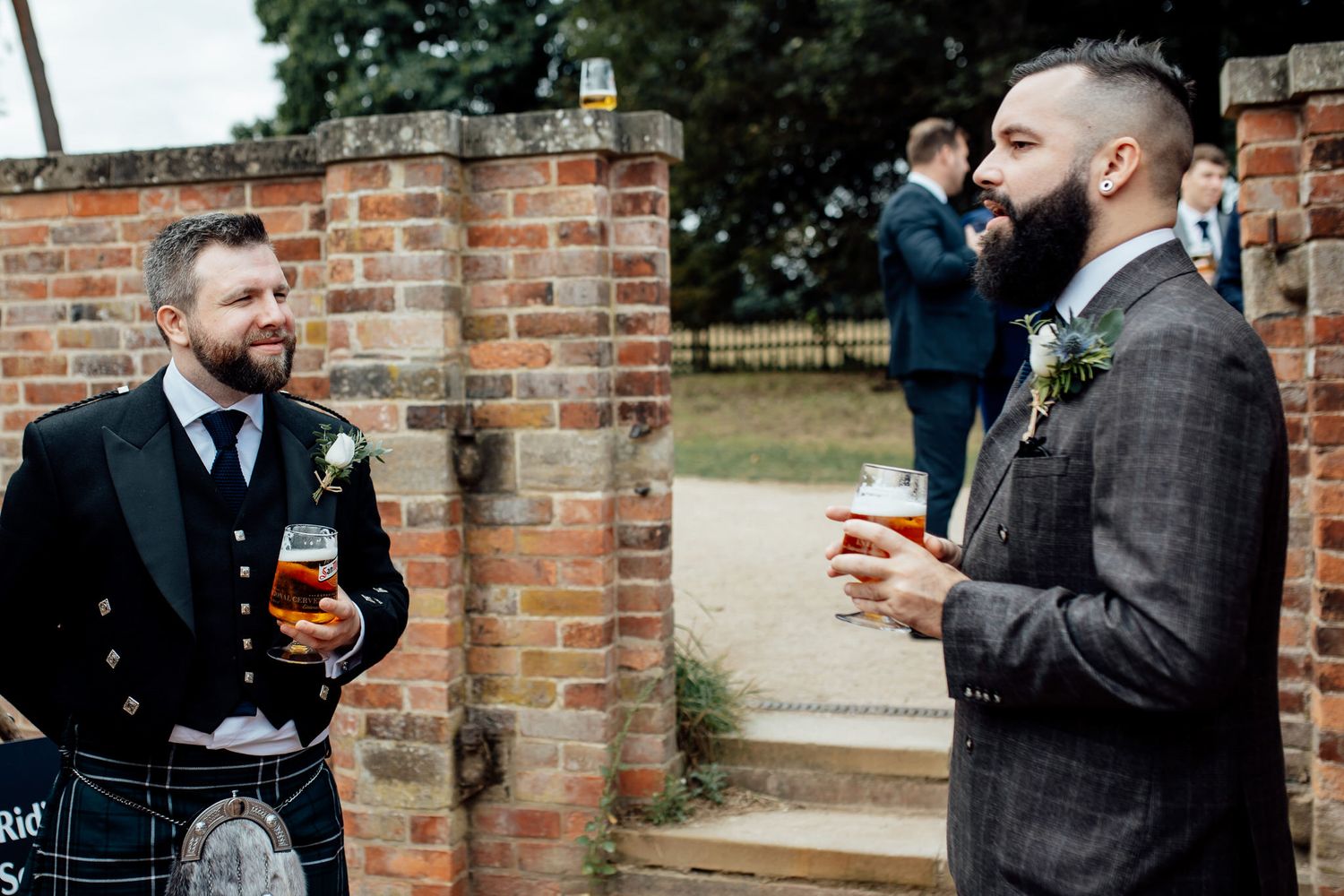 Two wedding guests in formal attire sharing drinks and conversation by brick wall.