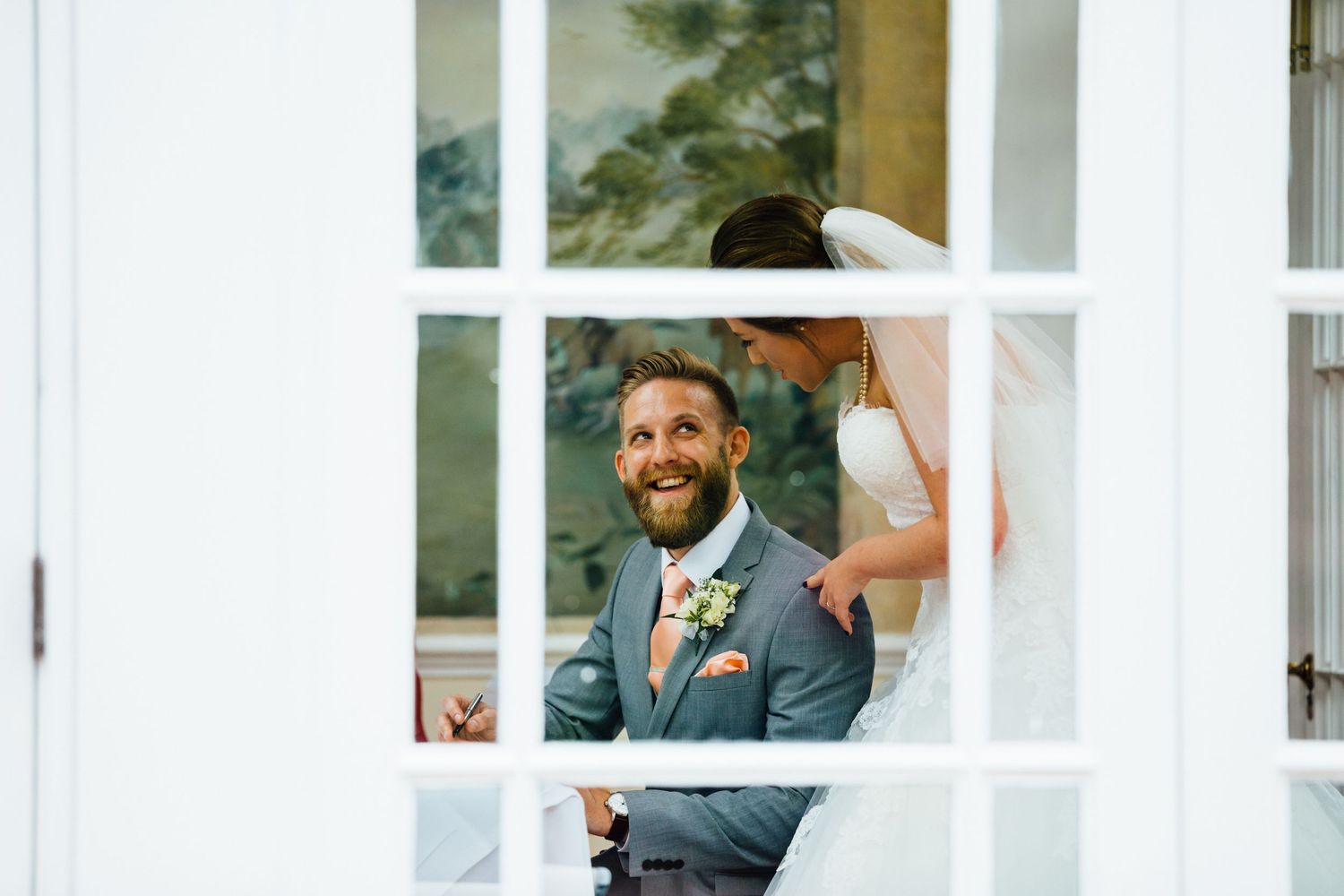 Wedding party poses in front of white windows and green foliage.