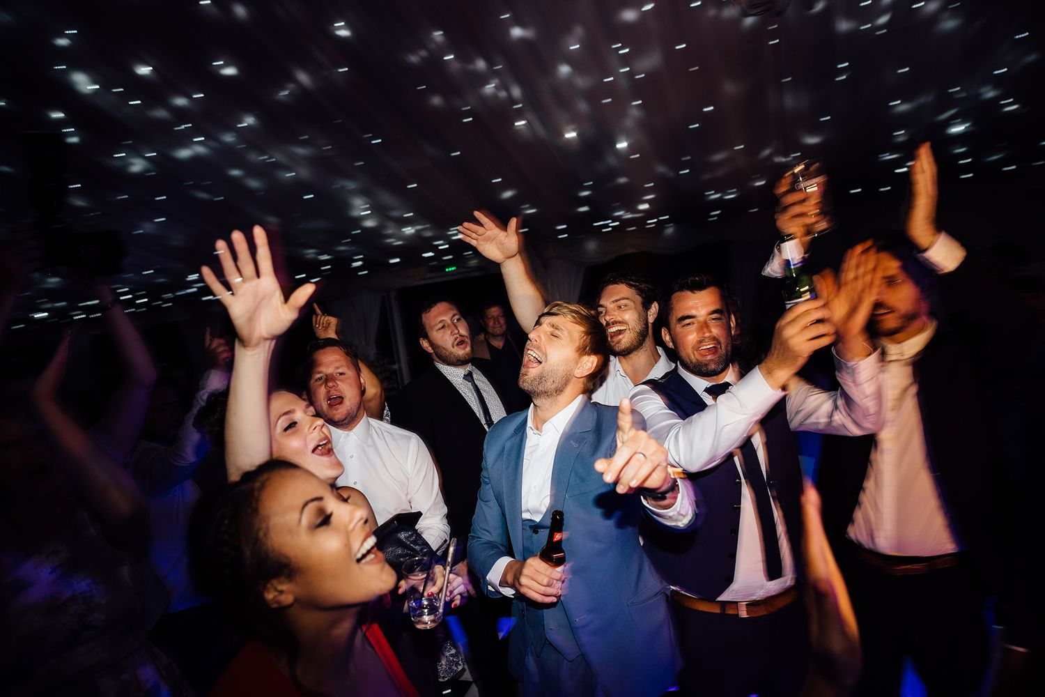 Wedding guests dance and celebrate with raised hands under sparkling lights at a reception party.