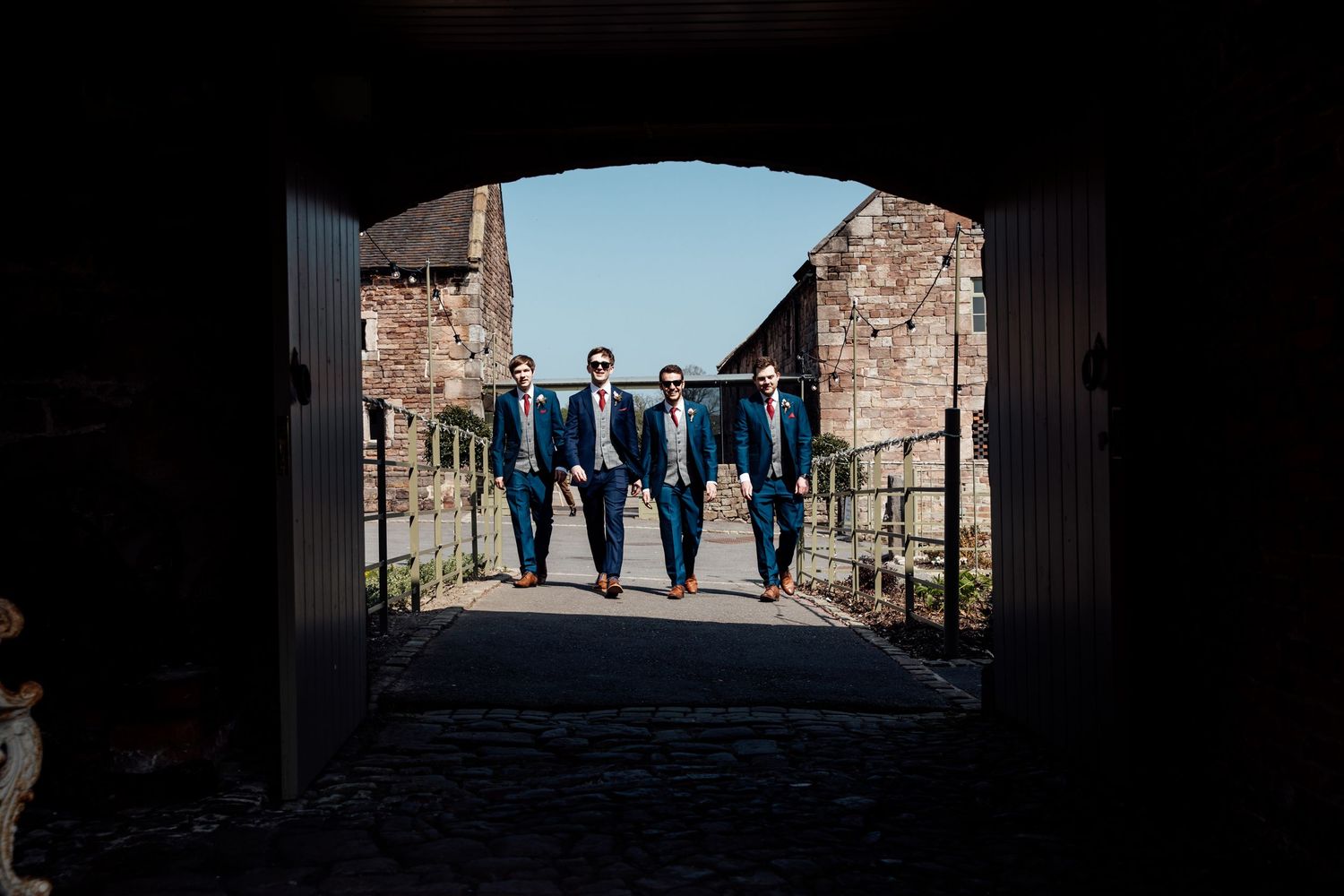Four groomsmen in navy suits walk through a stone archway at a historic venue.