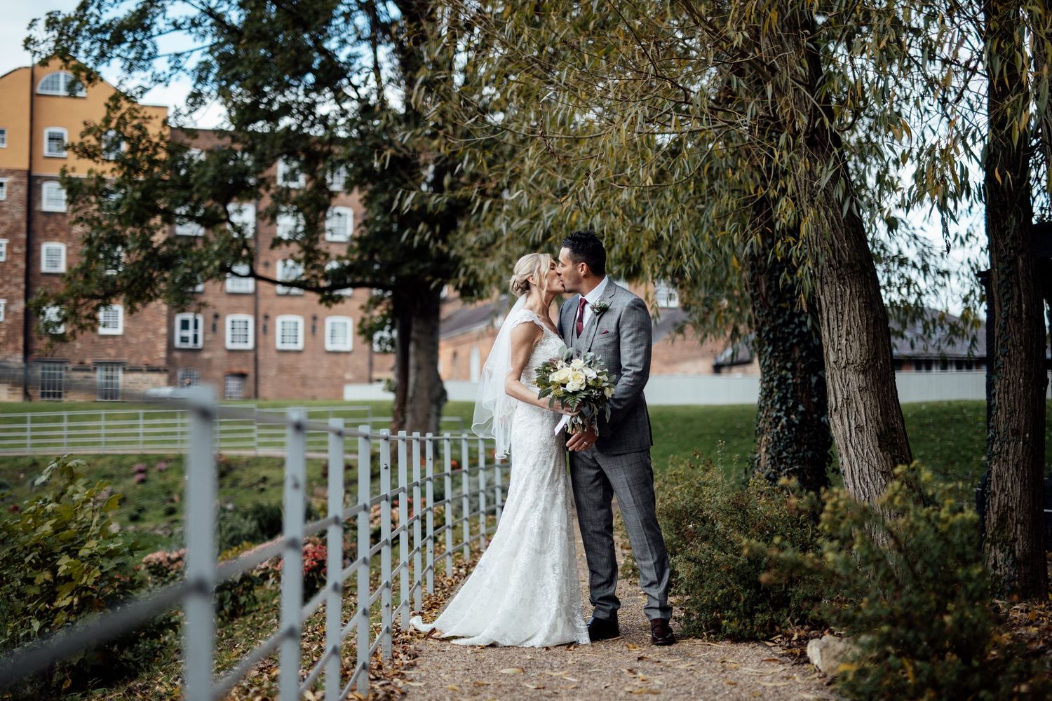 A couple in wedding attire shares a romantic moment under tall trees near a white fence at a rustic venue.