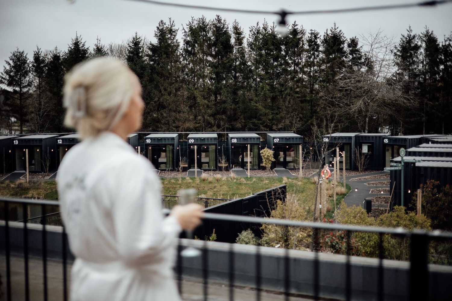 A woman in a white robe stands on a balcony overlooking rows of small cabins surrounded by trees.