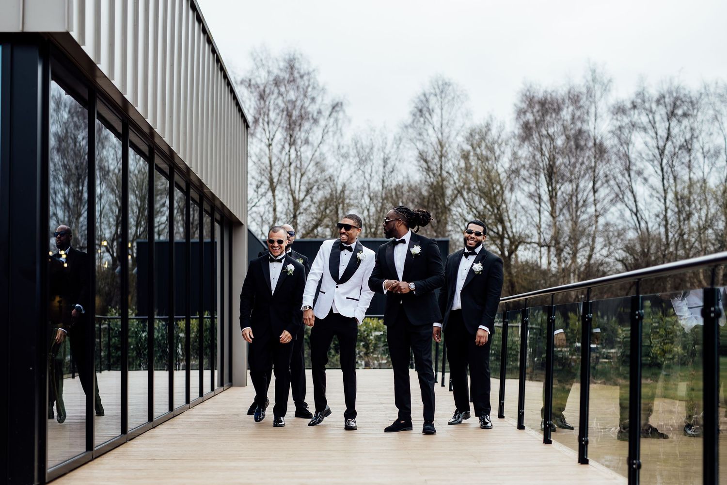 Groom in white tuxedo laughing with groomsmen in black suits on a modern outdoor terrace.