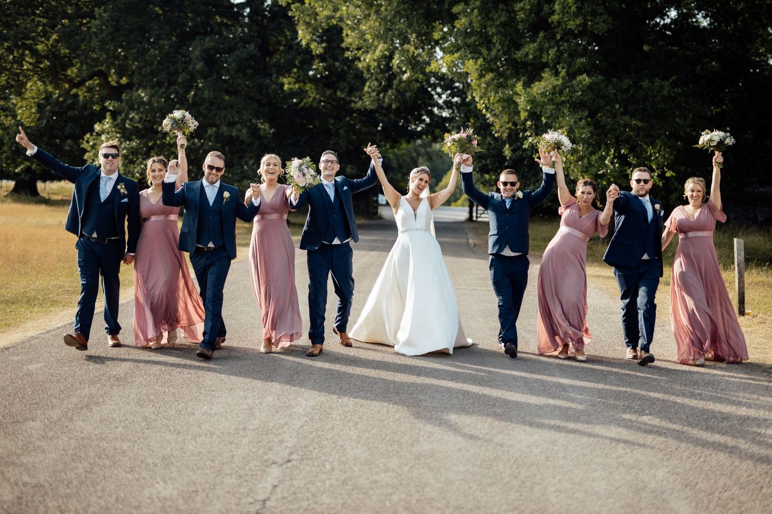 Wedding party celebrates on a path with bridesmaids in pink dresses and groomsmen in navy suits.