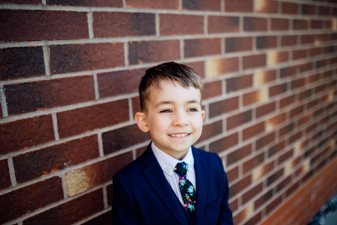 Young boy in navy suit smiles while standing against brick wall.