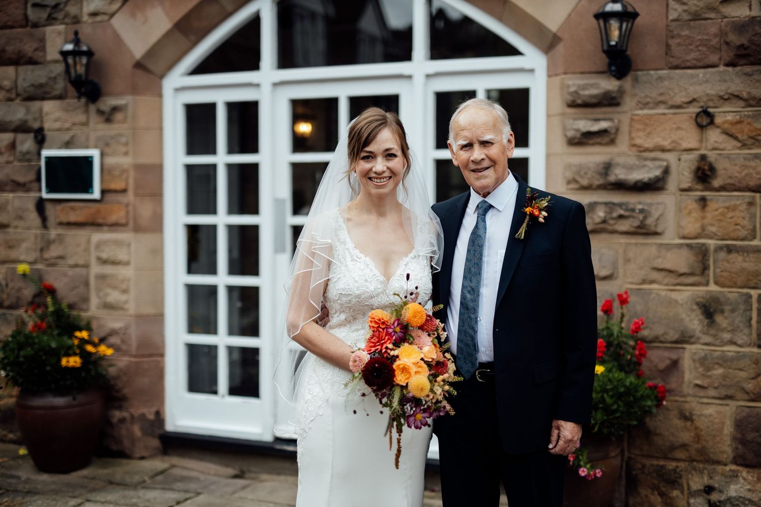 A bride in white lace gown holds vibrant autumn bouquet outside stone building entrance.