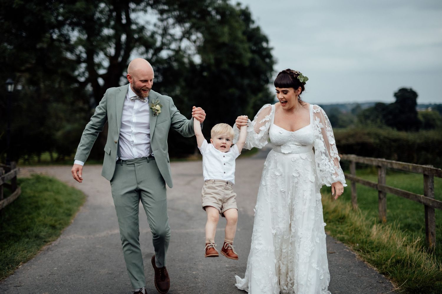 Wedding couple and child walking along a countryside path surrounded by green fields.