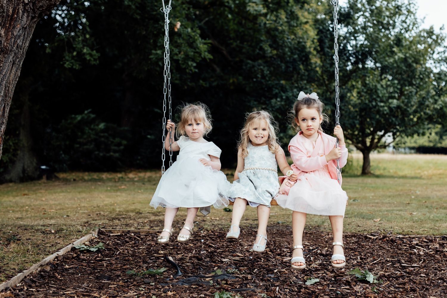 Children in white dresses sitting on tree stumps at an outdoor wedding.
