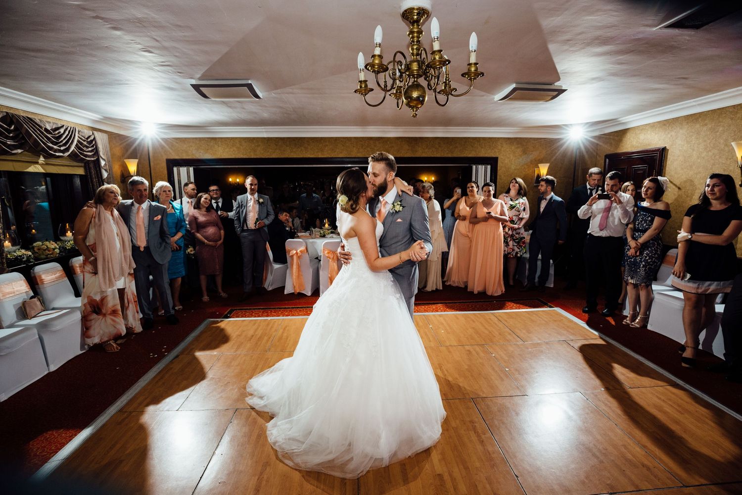 Wedding reception ballroom with chandelier and guests dancing on wooden floor.
