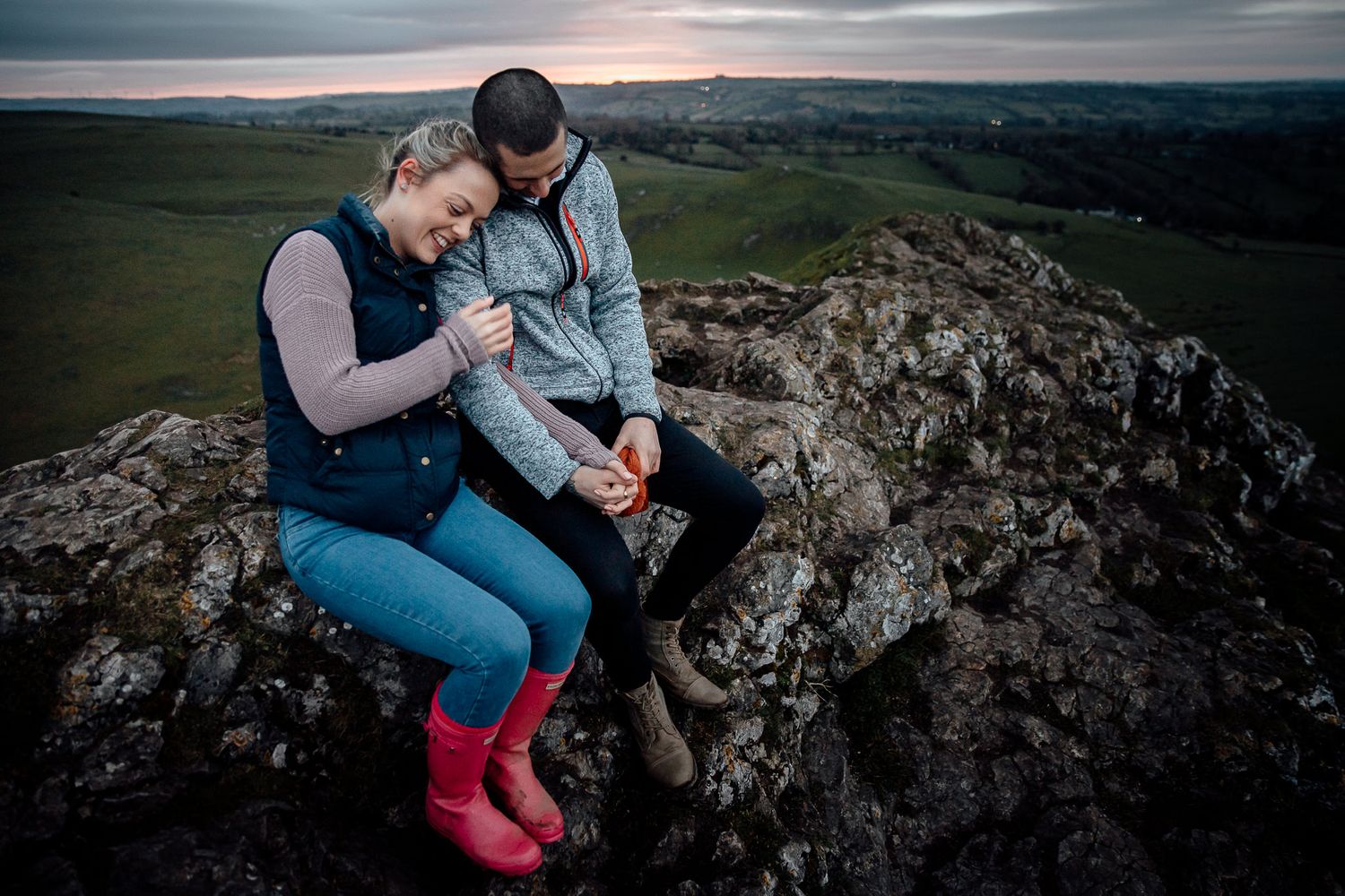 A couple sits intimately together on rocky cliffs overlooking a vast countryside landscape at sunset.