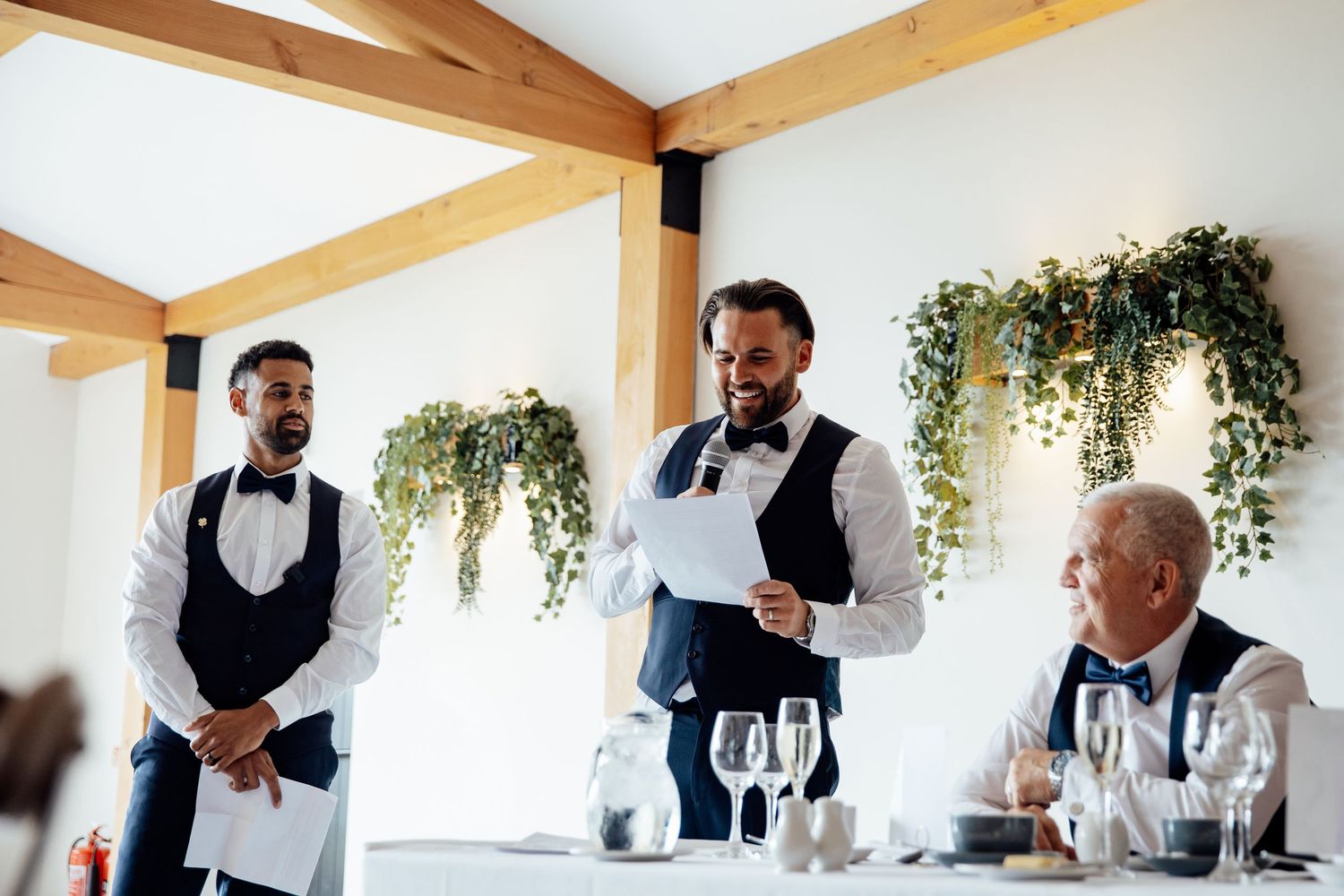 Wedding party members share joyful moments during indoor reception with wooden beam ceiling and hanging greenery.